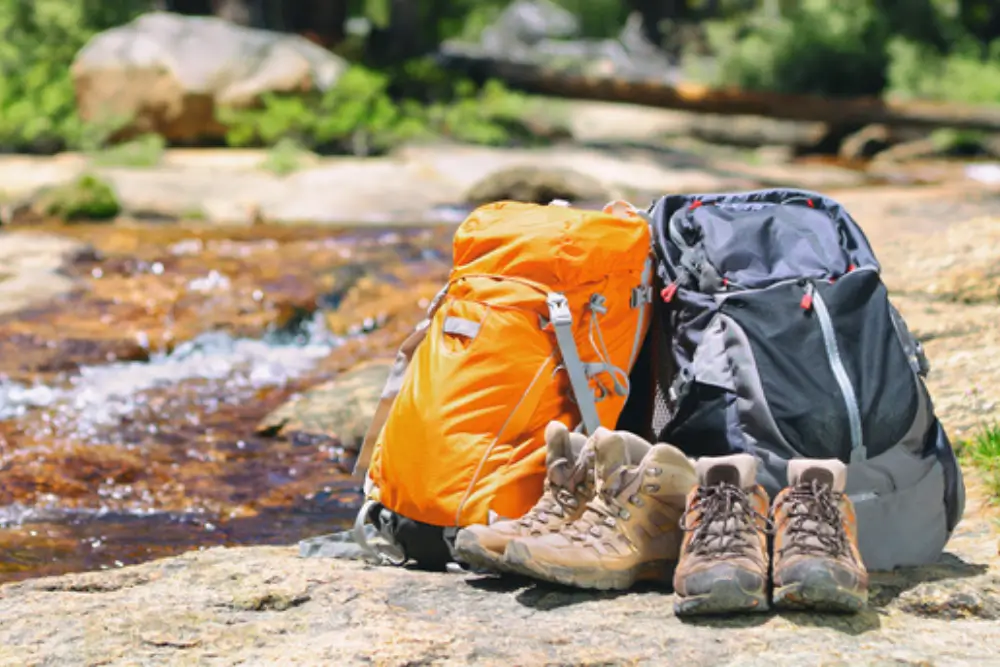 Outdoor gear like bags and shoes laid out beside flowing water on a hot summer day